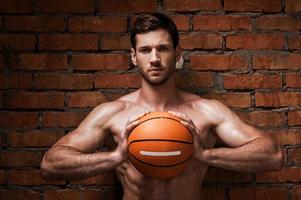 êtes-vous prêt à jouer confiant jeune homme musclé tenant un ballon de basket tout en se tenant contre le mur de briques photo
