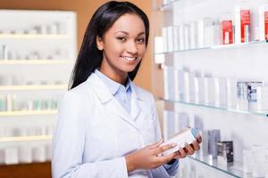 choix professionnel. belle jeune femme africaine en blouse de laboratoire tenant un récipient avec des médicaments et regardant la caméra avec le sourire tout en se tenant dans la pharmacie photo