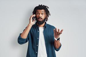 charmant jeune homme africain en tenue décontractée parlant au téléphone en se tenant debout sur fond gris photo