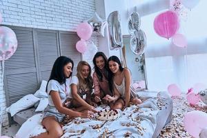 l'heure du gâteau. quatre belles jeunes femmes souriantes en pyjama mangeant du gâteau tout en organisant une soirée pyjama dans la chambre avec des ballons partout photo