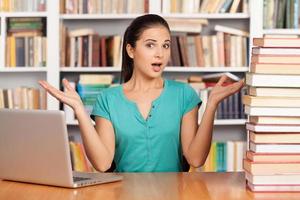 quoi choisir une jeune femme confuse assise au bureau de la bibliothèque avec une pile de livres et un ordinateur portable dessus photo