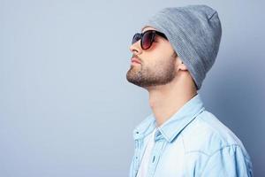 cool beau. vue latérale d'un beau jeune homme élégant en lunettes de soleil et chapeau regardant loin en se tenant debout sur fond gris photo