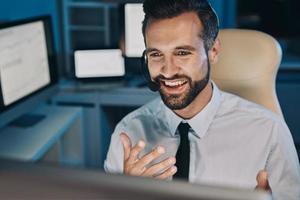 heureux jeune homme dans un casque regardant l'ordinateur et faisant des gestes tout en restant tard au bureau photo