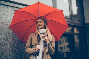 se sentir au chaud et protégé. jolie jeune femme souriante portant un parapluie et une tasse de café en se tenant debout dans la rue photo