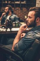 l'air génial comme jamais. vue latérale d'un beau jeune homme barbu regardant loin et gardant la main sur le menton tout en étant assis sur une chaise au salon de coiffure photo