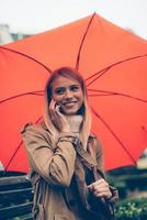 belle conversation avec un ami. jolie jeune femme souriante portant un parapluie et parlant au téléphone portable tout en étant assis sur le banc à l'extérieur photo