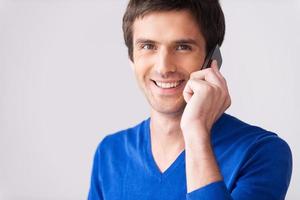 bonne conversation. beau jeune homme en pull bleu parlant au téléphone mobile et souriant en se tenant debout sur fond gris photo