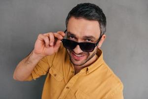 Hé bébé vue de dessus d'un beau jeune homme ajustant ses lunettes de soleil et souriant en se tenant debout sur fond gris photo