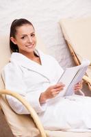 détente au centre de spa. belle jeune femme en peignoir se détendre assis sur une chaise et lire un magazine photo