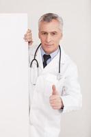 être en bonne santé médecin senior cheveux gris en uniforme regardant par l'espace de copie et faisant des gestes tout en étant isolé sur blanc photo