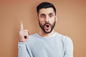 surpris jeune homme en tenue décontractée regardant la caméra et faisant une grimace en se tenant debout sur fond marron photo