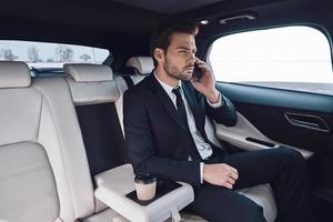 entreprise en mouvement. beau jeune homme en costume complet parlant au téléphone intelligent alors qu'il était assis dans la voiture photo