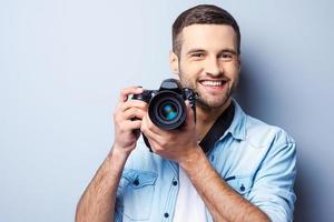 donne-moi un sourire beau jeune homme tenant un appareil photo numérique et souriant en se tenant debout sur fond gris