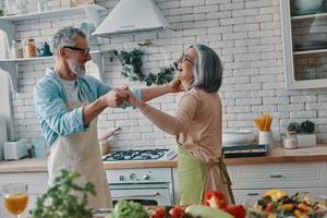 couple de personnes âgées ludique en tablier dansant et souriant tout en préparant un dîner sain à la maison photo