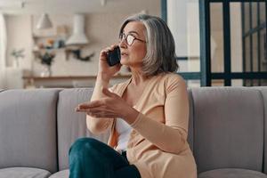 belle femme âgée en vêtements décontractés parlant au téléphone intelligent tout en étant assise sur le canapé à la maison photo