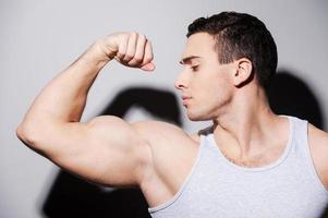 montrant son biceps parfait. beau jeune homme musclé montrant son biceps d'une part en se tenant debout sur fond gris photo