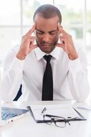 stressé et fatigué. jeune homme africain déprimé en tenue de soirée tenant la tête dans les mains et gardant les yeux fermés assis sur son lieu de travail photo