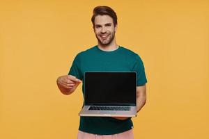 beau jeune homme souriant dans des vêtements décontractés portant un ordinateur portable et un moniteur de pointage photo