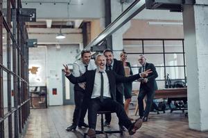 hommes d'affaires ludiques poussant leur patron sur la chaise de bureau tout en courant dans le couloir photo