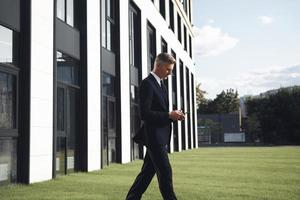 homme d'affaires mature confiant tenant un téléphone portable tout en marchant près d'un immeuble de bureaux photo