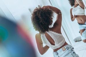 femme sensuelle. vue arrière de la belle jeune femme africaine gardant la main dans les cheveux tout en se tenant devant le miroir à la maison photo