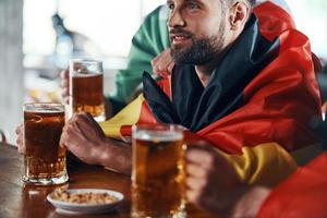 jeunes hommes couverts de drapeaux nationaux dégustant une bière tout en regardant un match de sport dans le pub photo