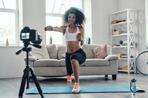 séduisante jeune femme africaine faisant de l'exercice en utilisant le poids de la main et souriant tout en faisant une vidéo sur les réseaux sociaux photo