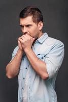 juste un instant pour tout repenser. vue latérale d'un beau jeune homme gardant les mains jointes et regardant pensif en se tenant debout sur fond gris photo