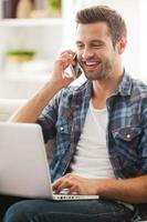 passer du temps en ligne. heureux jeune homme travaillant sur un ordinateur portable et parlant au téléphone assis sur un canapé photo