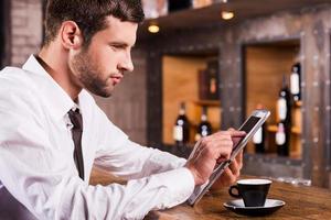 surfer sur le net au bar. vue latérale d'un beau jeune homme en chemise et cravate assis au comptoir du bar et travaillant sur une tablette numérique photo