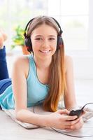 appréciant sa musique préférée. jolie adolescente au casque écoutant de la musique et regardant la caméra en position allongée sur le sol de son appartement photo