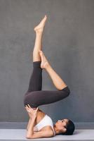 fort et gracieux. vue latérale d'une belle jeune femme africaine en vêtements de sport pratiquant le yoga sur un tapis d'exercice sur fond gris photo