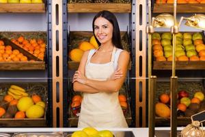 seuls les fruits les plus frais de notre magasin. belle jeune femme en tablier gardant les bras croisés et souriant tout en se tenant dans une épicerie avec une variété de fruits en arrière-plan photo