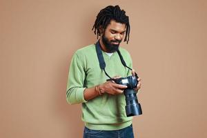 beau jeune photographe africain en vêtements décontractés à l'aide d'un appareil photo numérique et souriant en se tenant debout sur fond marron