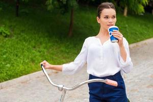 profiter de son temps libre dans le parc. jolie jeune femme buvant du café et regardant loin en marchant avec son vélo dans le parc photo
