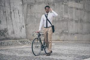 appréciant le mode de vie urbain. jeune homme confiant dans des verres regardant loin et tenant la main sur son vélo tout en marchant à l'extérieur photo