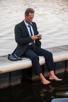 se sentir seul. homme d'affaires mature frustré regardant son téléphone portable tout en étant assis pieds nus sur le quai photo