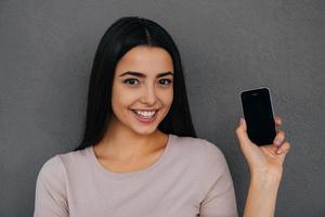 montrant son nouveau téléphone intelligent. belle jeune femme montrant son téléphone intelligent et souriant en se tenant debout sur fond gris photo