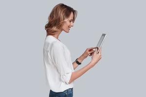 utilisant les technologies modernes. séduisante jeune femme tenant une tablette numérique et la regardant en se tenant debout sur fond gris photo