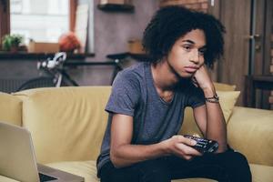 rien d'intéressant à regarder. jeune homme africain tenant une télécommande et ayant l'air ennuyé en regardant la télévision sur le canapé à la maison photo