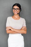 prêt à vous aider. confiante jeune femme service à la clientèle en gardant les bras croisés et en regardant la caméra en se tenant debout sur fond gris photo