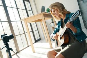 joyeuse jeune femme souriante et jouant de la guitare tout en jouant de la guitare devant un appareil photo numérique