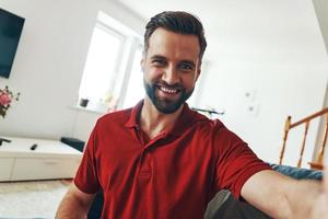 beau jeune homme en vêtements décontractés regardant la caméra et souriant tout en passant du temps à l'intérieur photo