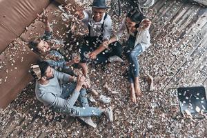 insouciant et heureux. vue de dessus de jeunes gens modernes buvant du champagne et souriant assis sur le sol avec des confettis volant autour photo