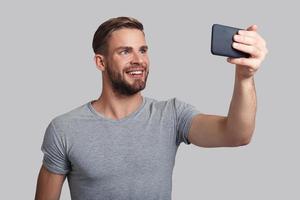 photo pour sa petite amie. beau jeune homme souriant prenant selfie en se tenant debout sur fond gris