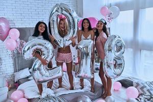 la fête est sur quatre jolies jeunes femmes souriantes en pyjama tenant des ballons debout dans la chambre à la maison photo