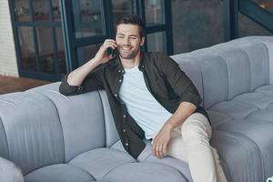 beau jeune homme parlant au téléphone intelligent et regardant ailleurs assis sur le canapé à la maison photo