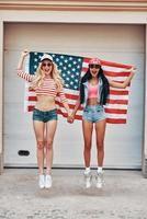 style américain. deux jeunes femmes gaies tenant un drapeau américain derrière elles tout en sautant contre la porte du garage photo