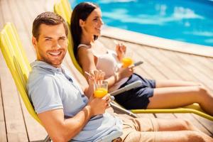 profitant de leur temps libre ensemble. vue latérale d'un beau jeune couple assis sur les chaises longues au bord de la piscine et buvant des cocktails pendant que l'homme regarde la caméra et sourit photo