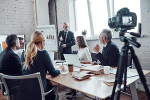 une équipe commerciale prospère écoute un homme qui fait une présentation tout en filmant une réunion du personnel dans la salle du conseil photo